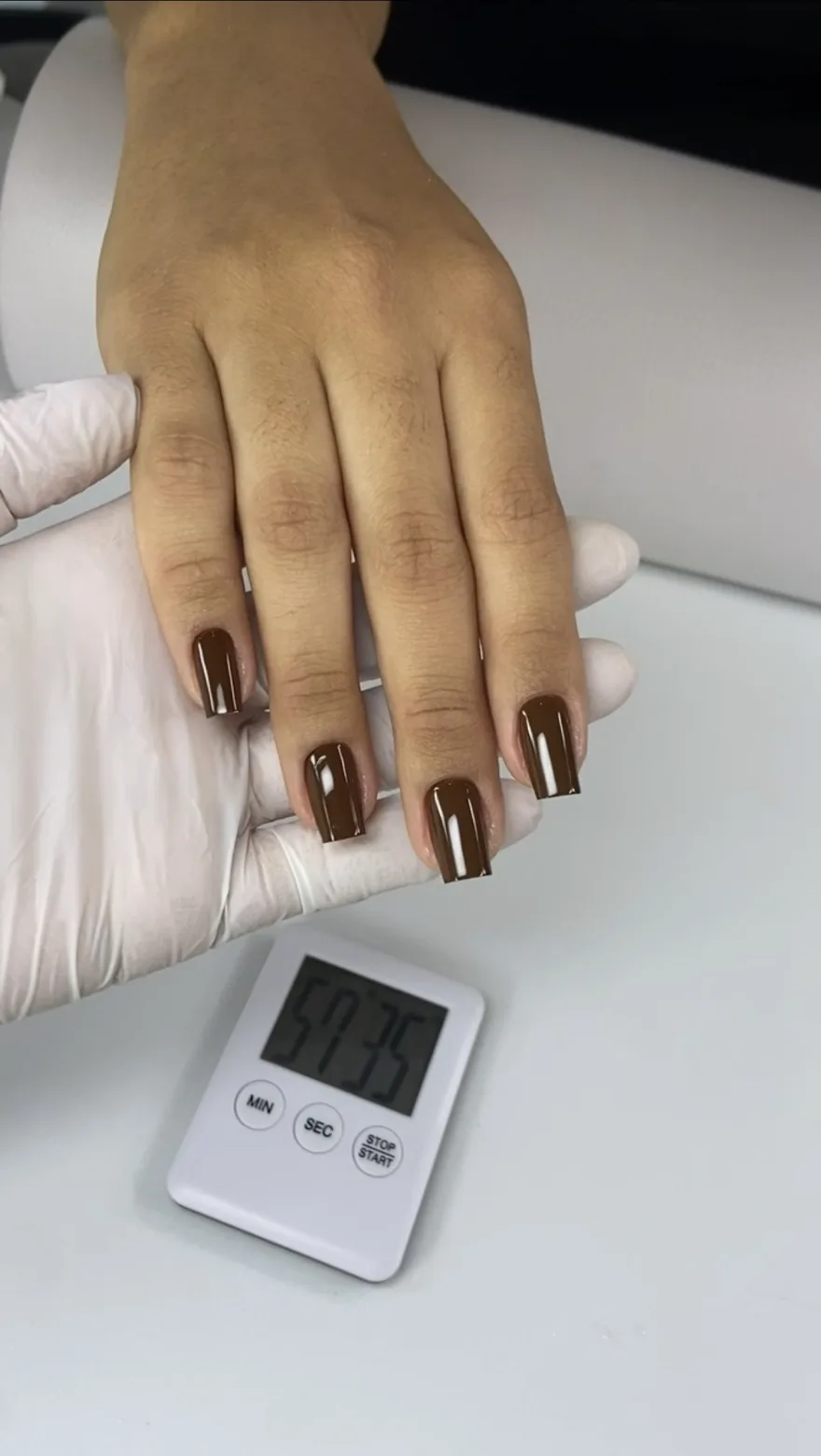 Mão de cliente com unhas marrons prontas sendo segurada por mão enluvada, com um cronômetro marcando o tempo