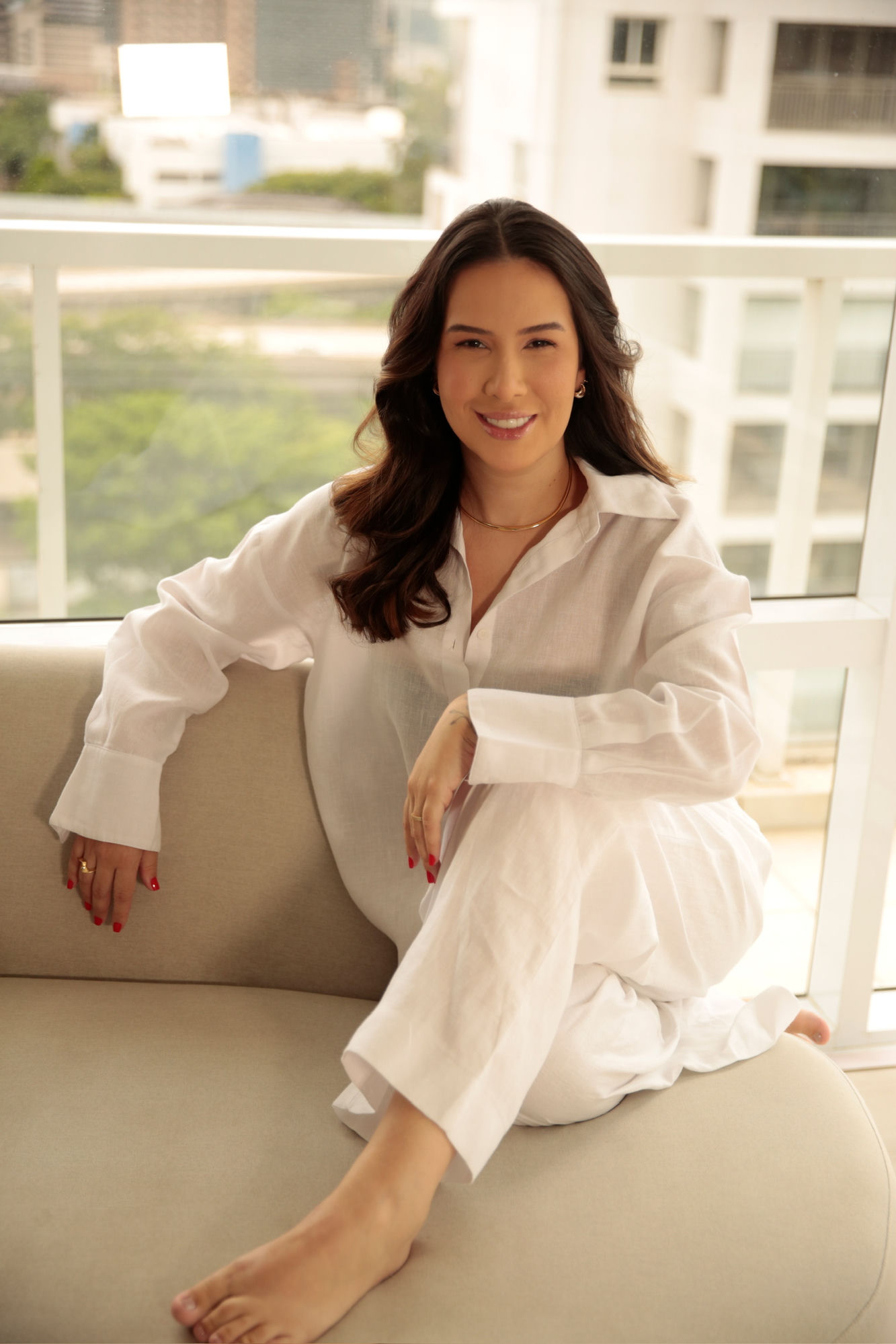 Thais Bessa sorrindo para a camera, de corpo inteiro, com look branco e luz natural, sentada de forma relaxada