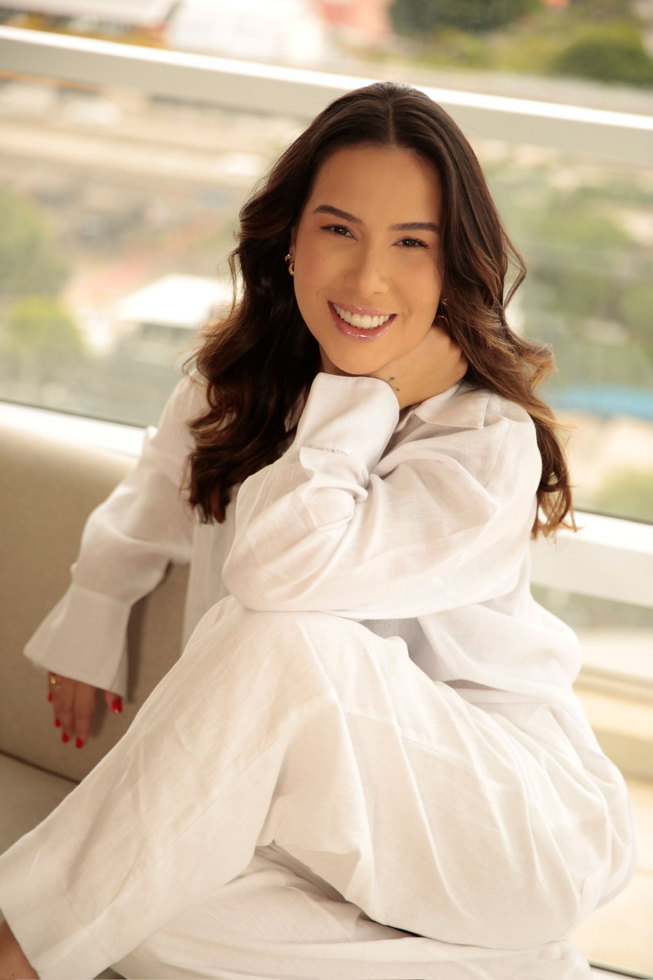 Thais Bessa, nail designer e instrutora, sorrindo para a camera, sentada e de look branco com luz natural